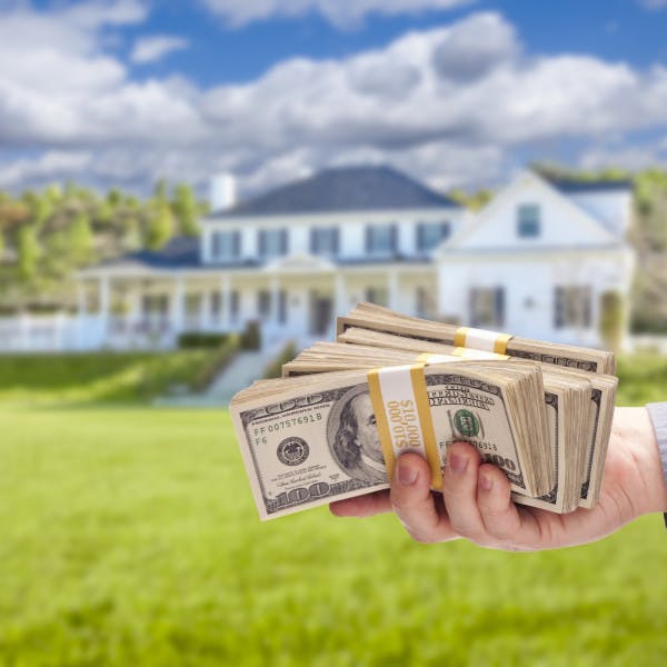 A businessman holds a wad of hundred-dollar bills in front of a luxury home