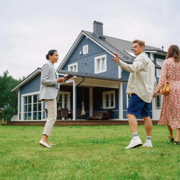 A real estate agent walks with a young couple through a large yard outside a suburban home.