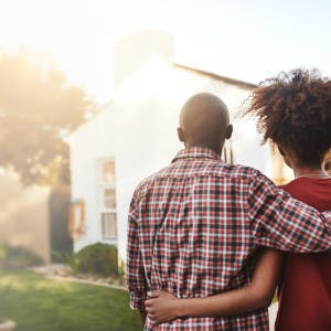 A Black couple stands outside with their arms around each other looking at a house
