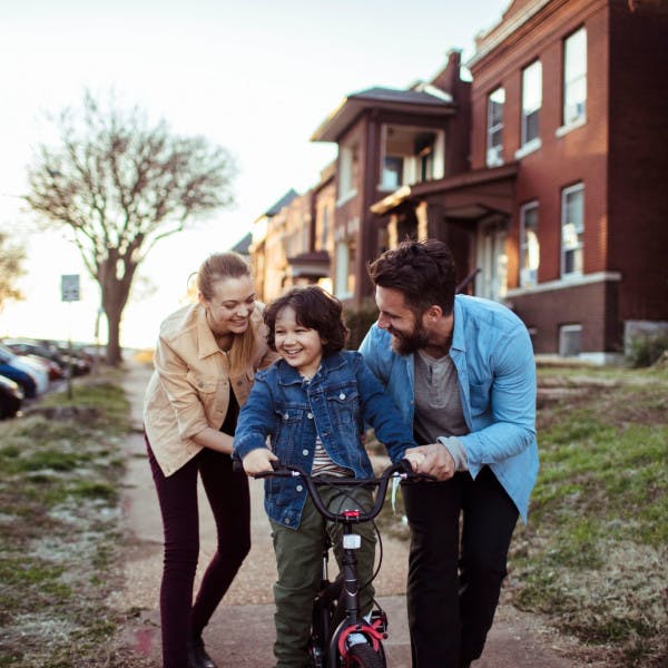 A young couple helps their child learn to ride a bike along the sidewalk