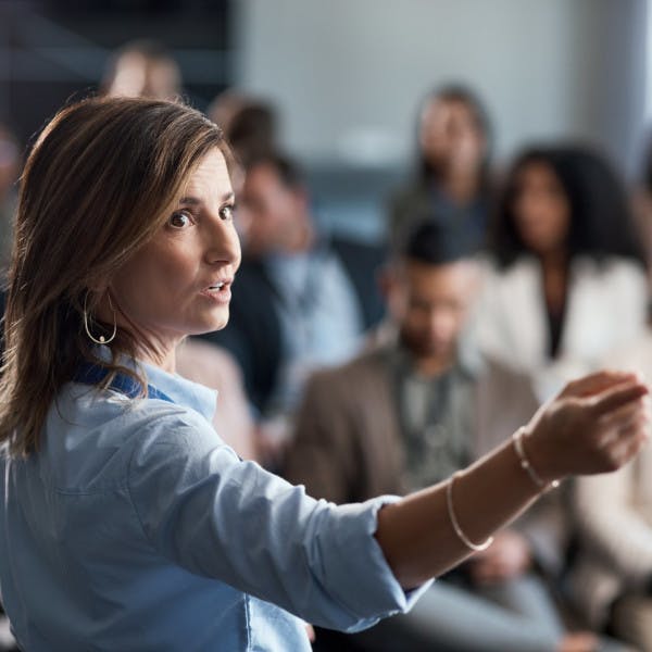 A woman business trainer points to a presentation while speaking to a group of businesspeople